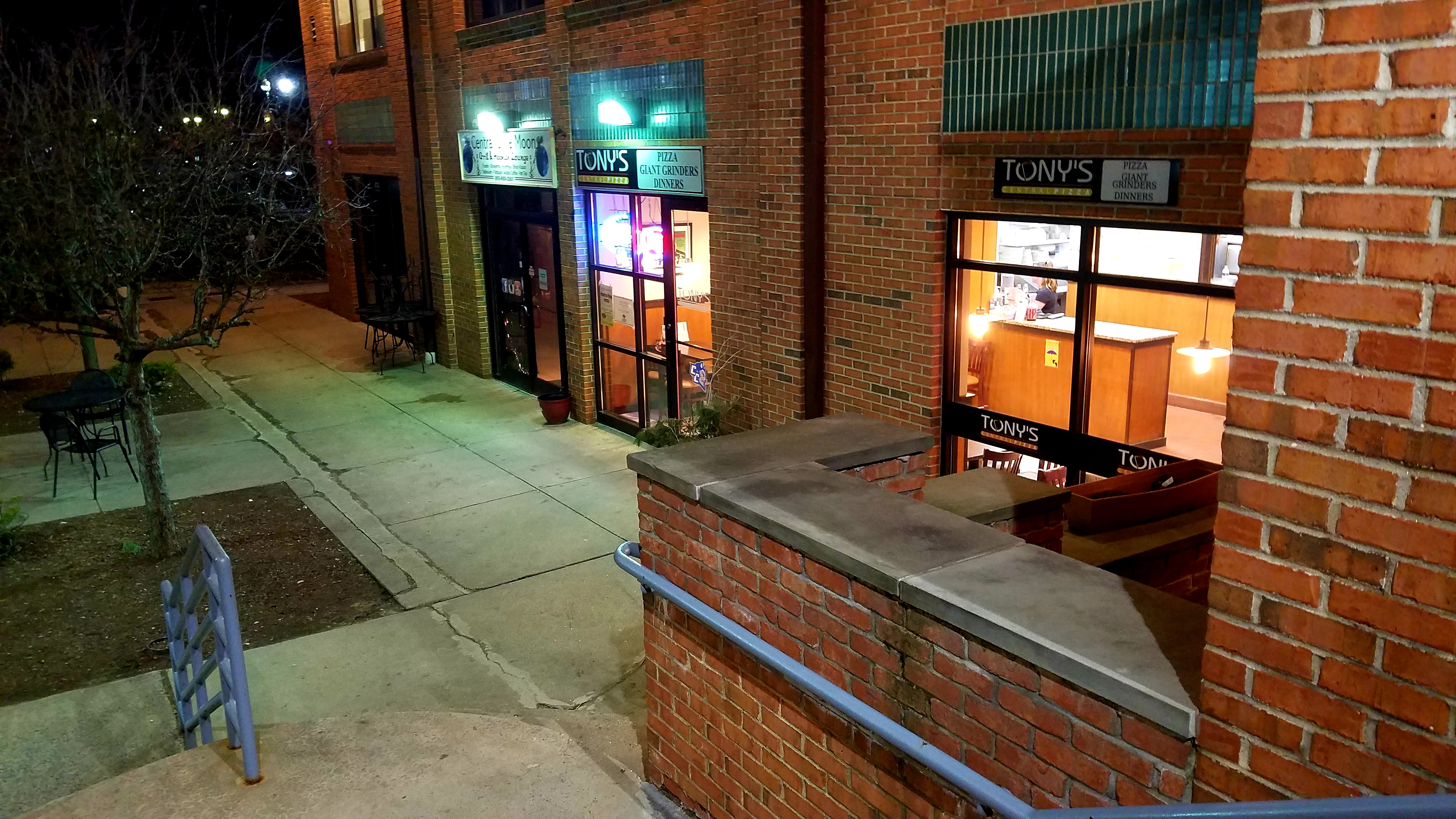 Exterior storefront of Tony's Central Pizza at night, showing the brick facade, illuminated windows, and signage identifying the business location.
