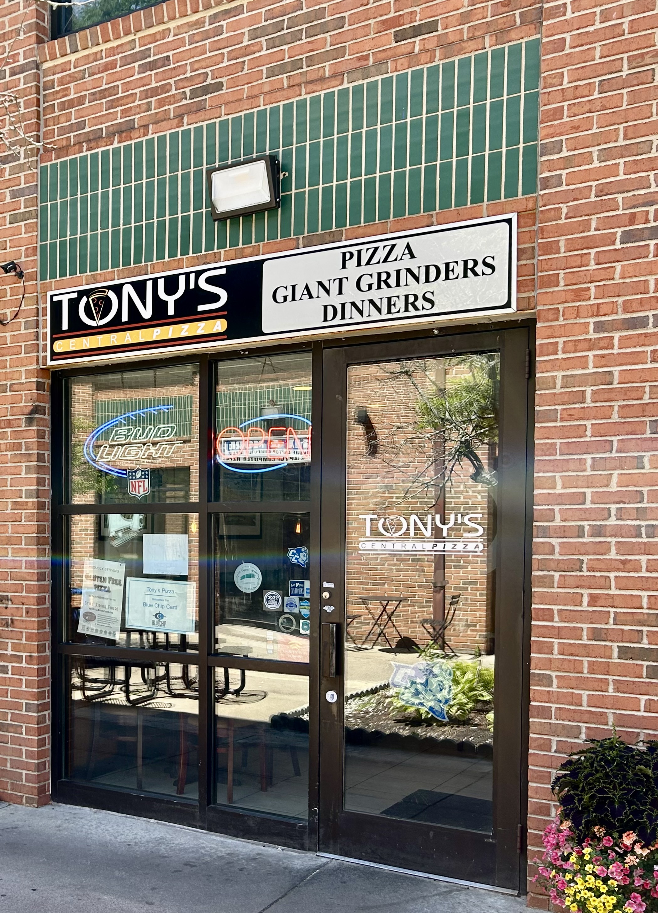 Storefront entrance with Tony's logo and signage reading 'PIZZA GIANT GRINDERS DINNERS' on the brick facade, showing glass doors and windows.