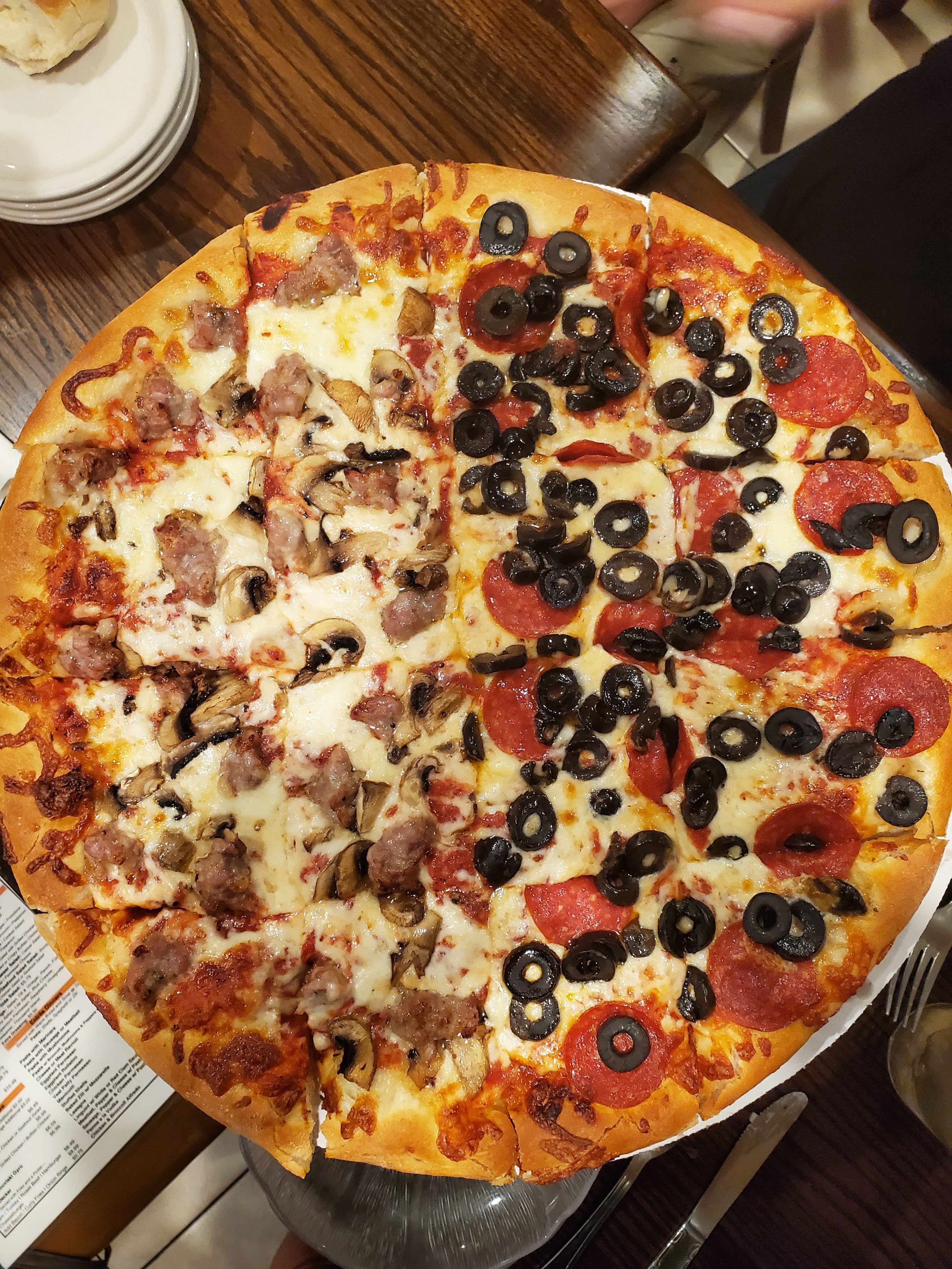 A whole pizza topped with meat, pepperoni, black olives, and mushrooms served on a white plate over a wooden table.