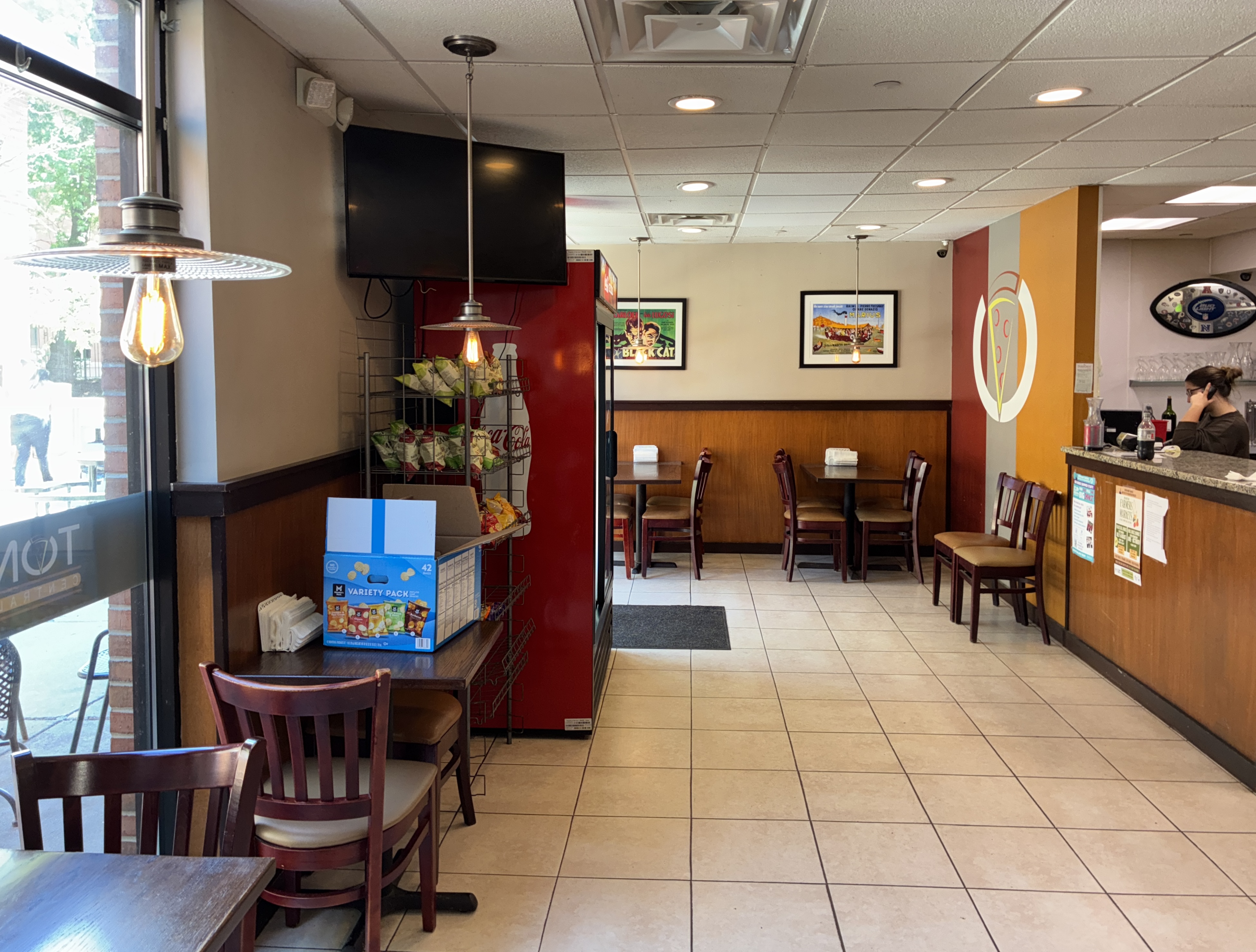 Interior dining room with wooden wainscoting, red and yellow accent walls, ceiling-mounted TV, tables with place settings, and counter area with staff visible.