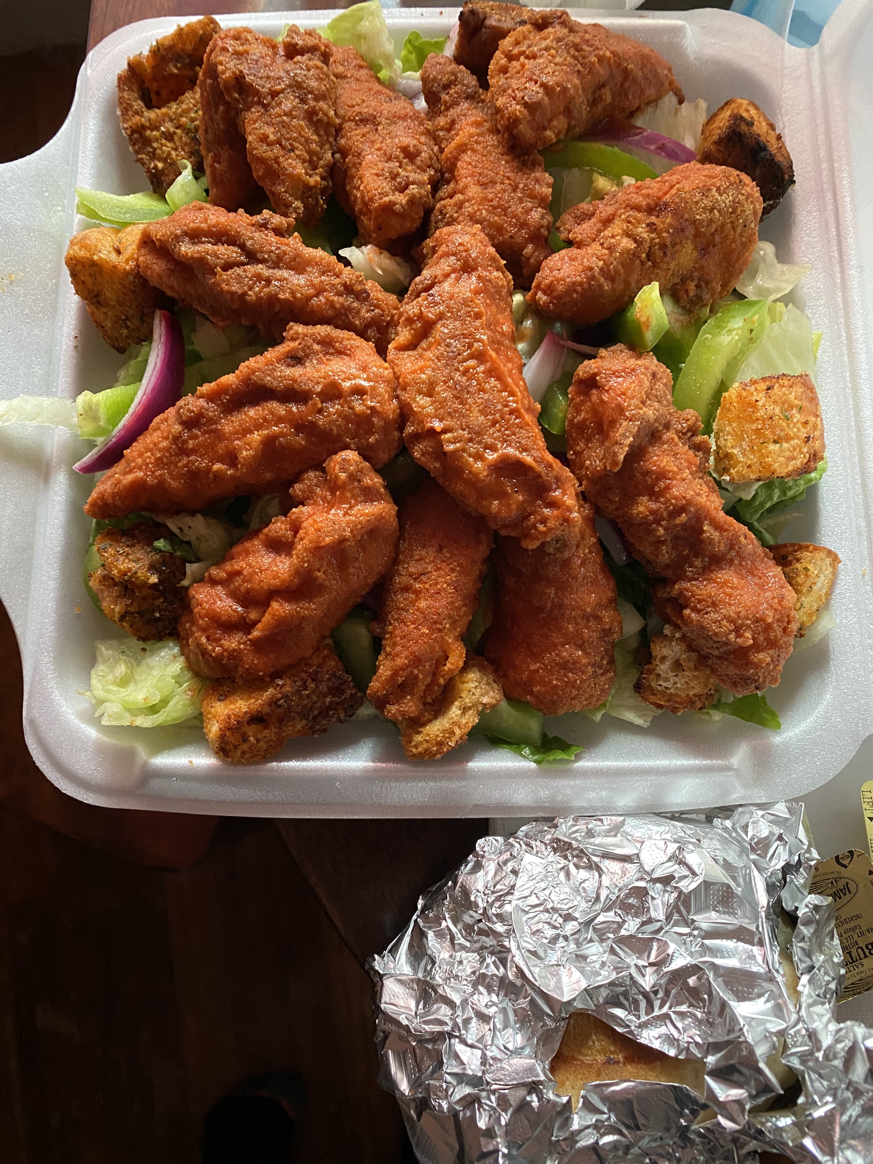 Fried chicken wings in a takeout container lined with lettuce, accompanied by a foil-wrapped side item.