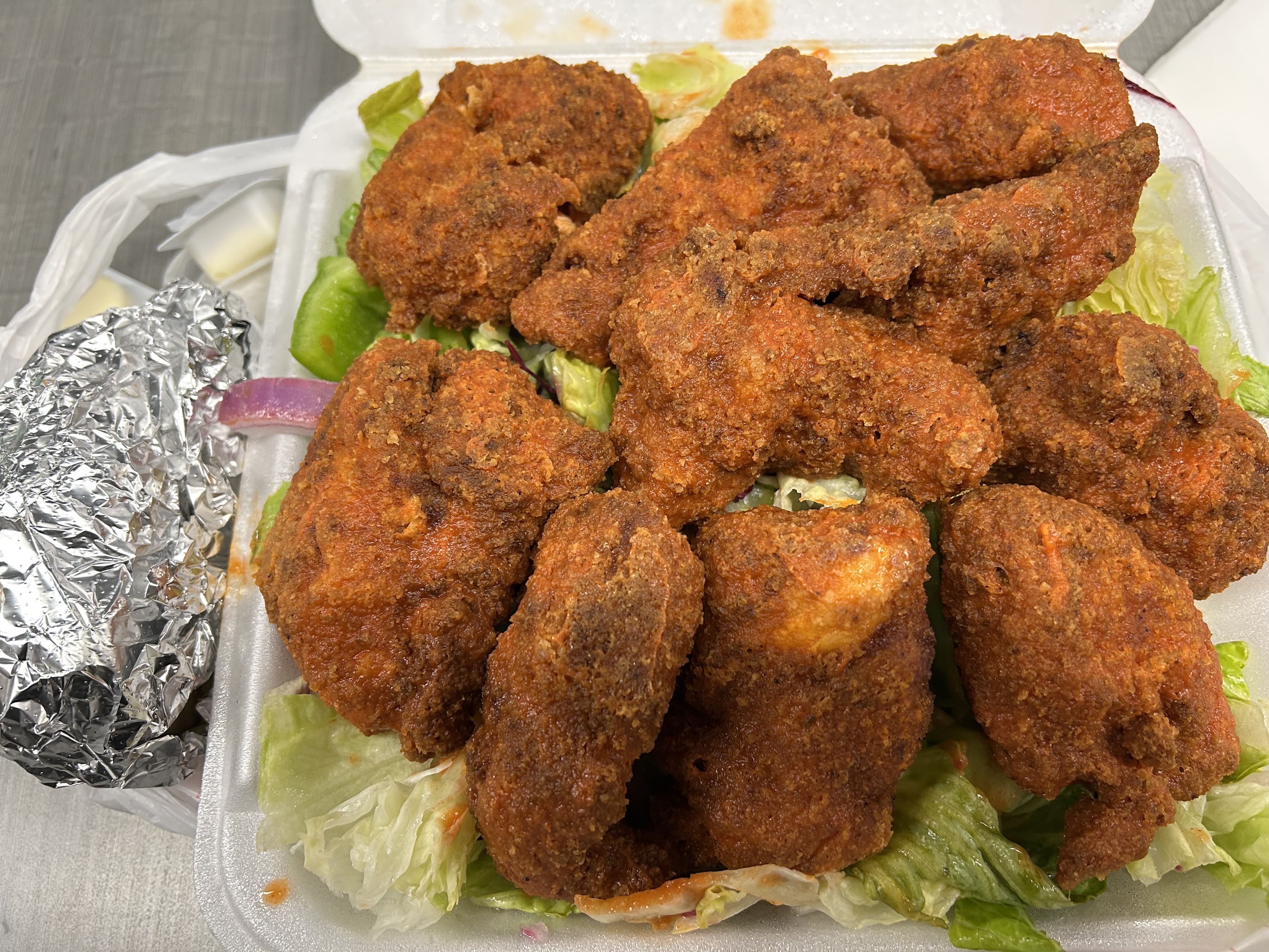 Fried chicken wings in a takeout container on lettuce with a foil-wrapped side and dipping container visible.