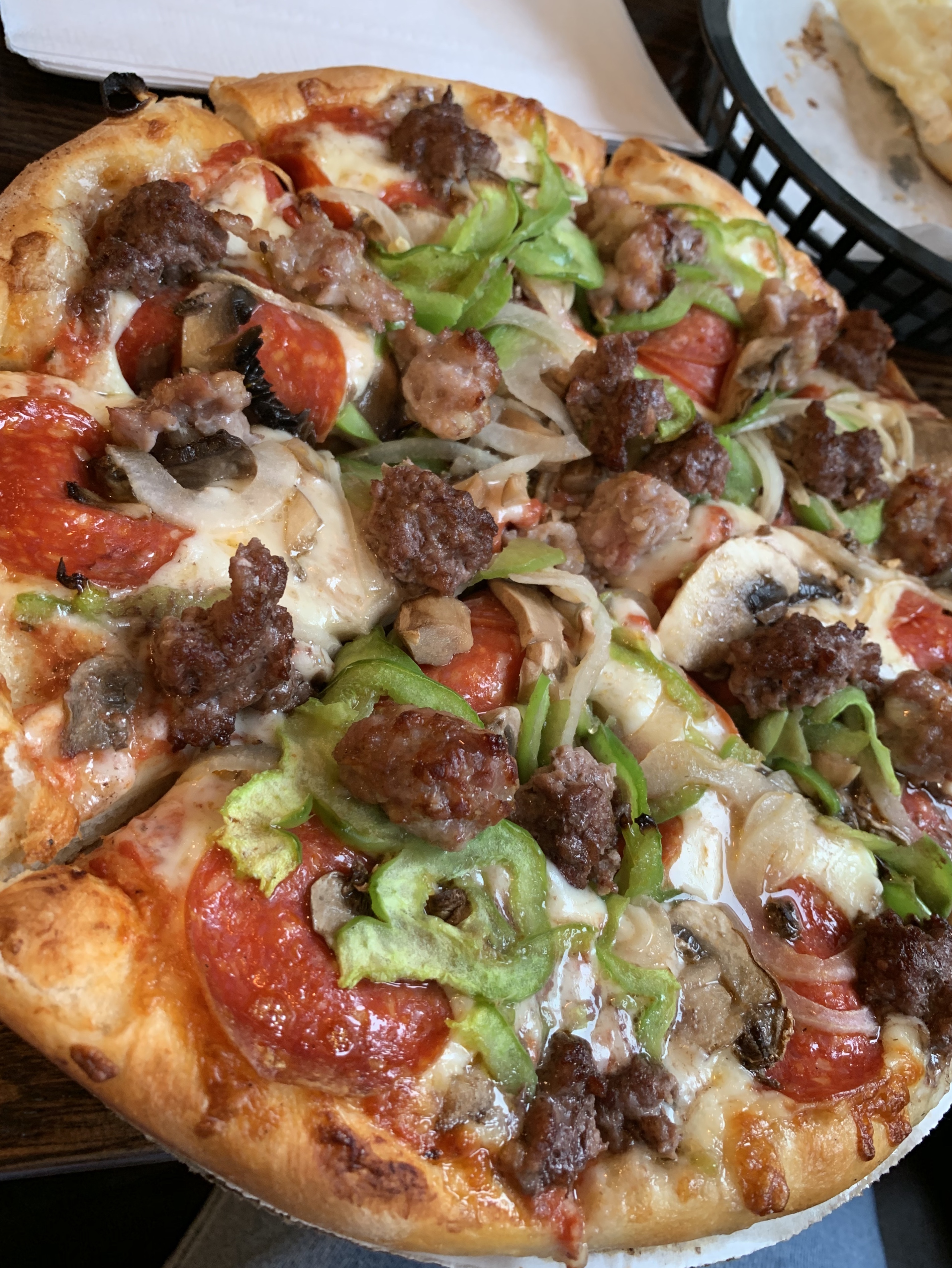 A pizza topped with sausage, pepperoni, onions, mushrooms, and green peppers, served in a paper-lined basket.