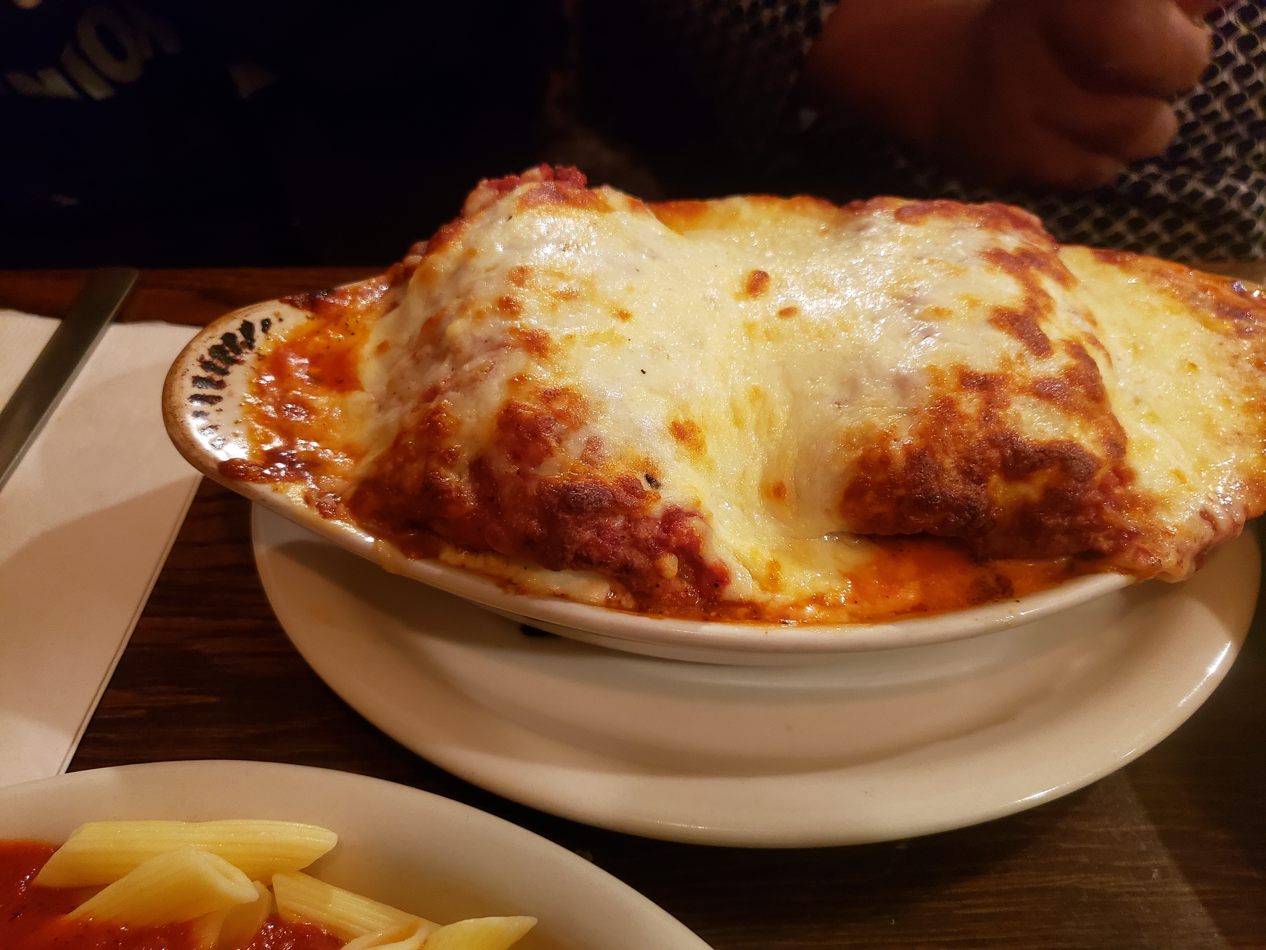 A baked pasta dish in a white ceramic bowl topped with melted cheese and red sauce, with fries visible at the plate edge.